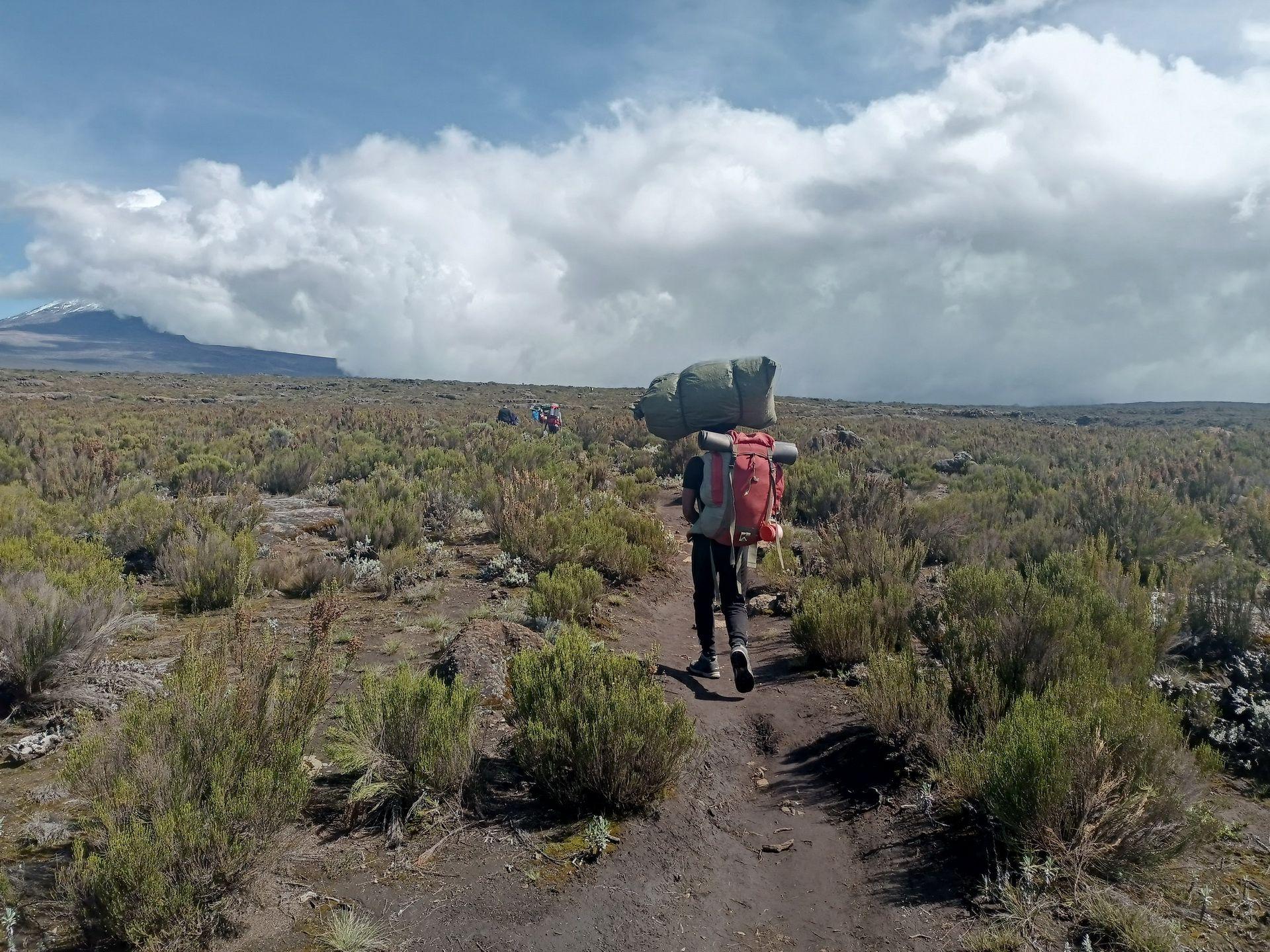 A porter carrying a load across low moorland under a big sky
